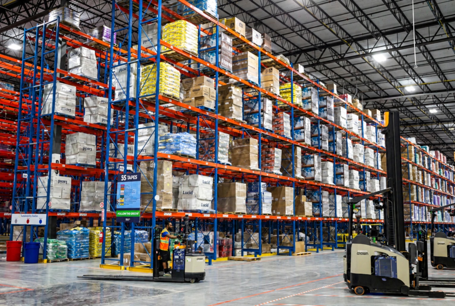 Stocked warehouse pallet racks with reach trucks, a lift operator and marked floor lanes