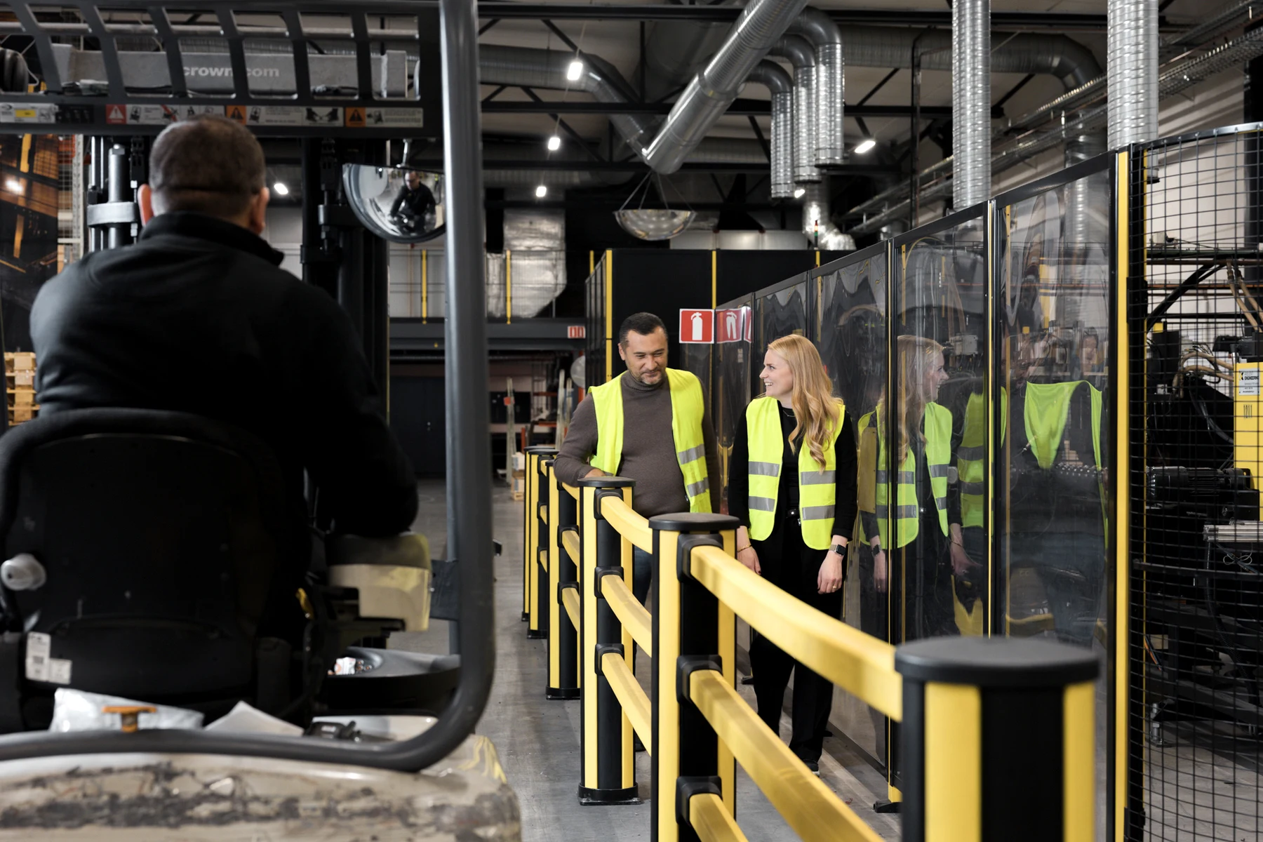 Two pedestrians behind yellow industrial railing with a forklift in the separate vehicle aisle